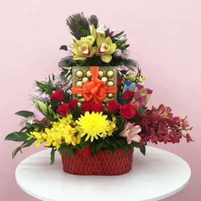 A decorative basket filled with colorful flowers and assorted chocolates placed on a wooden table.