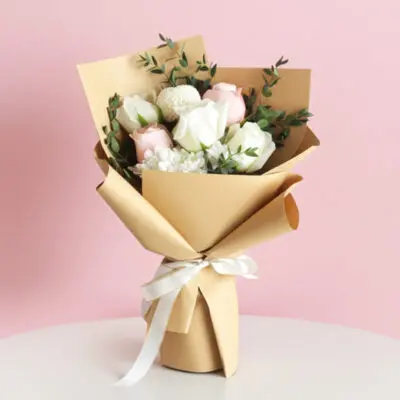 A hand bouquet featuring five white and pink roses arranged in a brown paper bag, alongside a ping pong and a pompom.