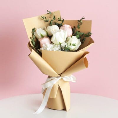 A hand bouquet featuring five white and pink roses arranged in a brown paper bag, alongside a ping pong and a pompom.