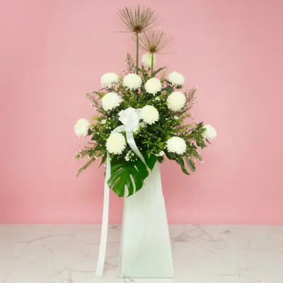 A white stand containing funeral flowers filled with delicate white flowers and green leaves.