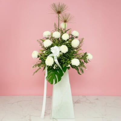 A white stand containing funeral flowers filled with delicate white flowers and green leaves.