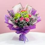 A bouquet featuring purple and pink carnations, button chrysanthemums, and baby's breath, elegantly wrapped in purple.