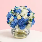 A floral arrangement featuring 3 blue hydrangeas, 1 white hydrangea, 4 blue orchids, 4 roses, and 5 wax flowers with greenery.