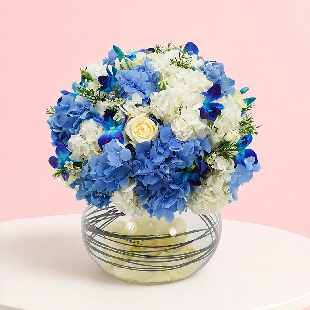 dazzling-hydrangea-with-orchid-in-fish-bowl_2026 A floral arrangement featuring 3 blue hydrangeas, 1 white hydrangea, 4 blue orchids, 4 roses, and 5 wax flowers with greenery.