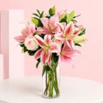 A clear vase holds pink stargazer lilies and light pink roses, creating an elegant floral arrangement.