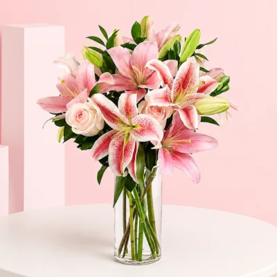 A clear vase holds pink stargazer lilies and light pink roses, creating an elegant floral arrangement.