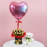 A teddy bear sits beside a heart-shaped balloon, both placed in a decorative vase.