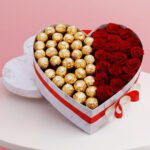A heart-shaped box filled with assorted chocolates and surrounded by fresh red roses.