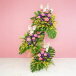 A Large Bouquet Of Mathilo, Gerbera, Eustoma, And Golden Shower Flowers Set Against A Pink Backdrop.
