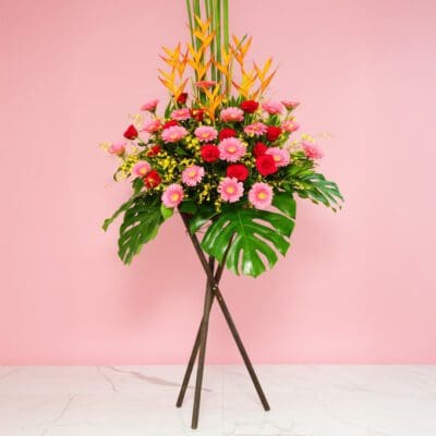 Colorful Flower Arrangement With Heliconia, Gerberas, And Roses Displayed On A Stand In Front Of A Pink Wall.