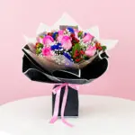 A hand bouquet of pink and blue roses with baby breath and cherry, presented in a black paper bag.