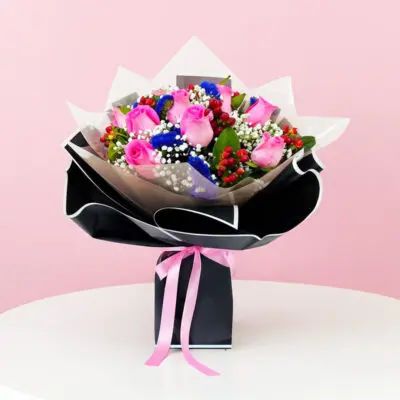A hand bouquet of pink and blue roses with baby breath and cherry, presented in a black paper bag.