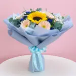 A hand bouquet featuring three sunflowers, pink and purple gerbera, baby breaths, and seasonal greens, tied with a blue ribbon.