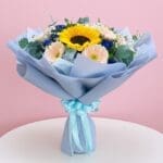 A hand bouquet featuring three sunflowers, pink and purple gerbera, baby breaths, and seasonal greens, tied with a blue ribbon.