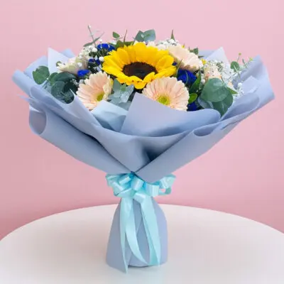 A hand bouquet featuring three sunflowers, pink and purple gerbera, baby breaths, and seasonal greens, tied with a blue ribbon.