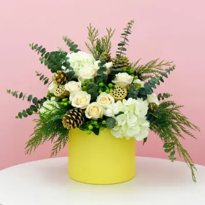 A yellow vase filled with white flowers and pine cones, placed beside a festive Christmas box.