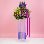 A grand opening stand vase filled with assorted flowers, including Gerbera and Matthiola, against a pink background.