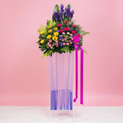 A grand opening stand vase filled with assorted flowers, including Gerbera and Matthiola, against a pink background.