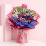 Hand bouquet of seasonal flowers in pastel shades, including lilies and spray roses, wrapped in vibrant pink paper.