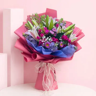 Hand bouquet of seasonal flowers in pastel shades, including lilies and spray roses, wrapped in vibrant pink paper.