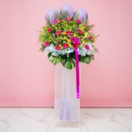 A grand opening flower stand featuring a vibrant arrangement of purple and pink Gerbera, orchids, and pompom flowers with balloons.