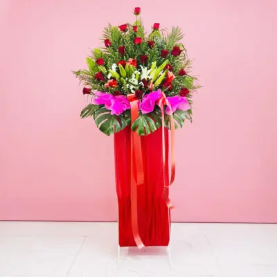 A red Grand Opening Stand filled with assorted flowers, including roses and lilies, adorned with a ribbon against a pink background.