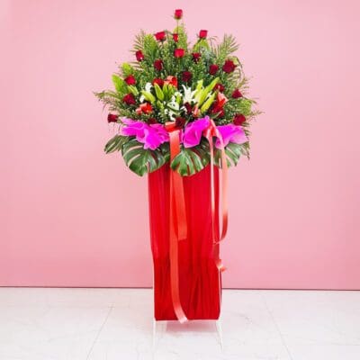 A red Grand Opening Stand filled with assorted flowers, including roses and lilies, adorned with a ribbon against a pink background.
