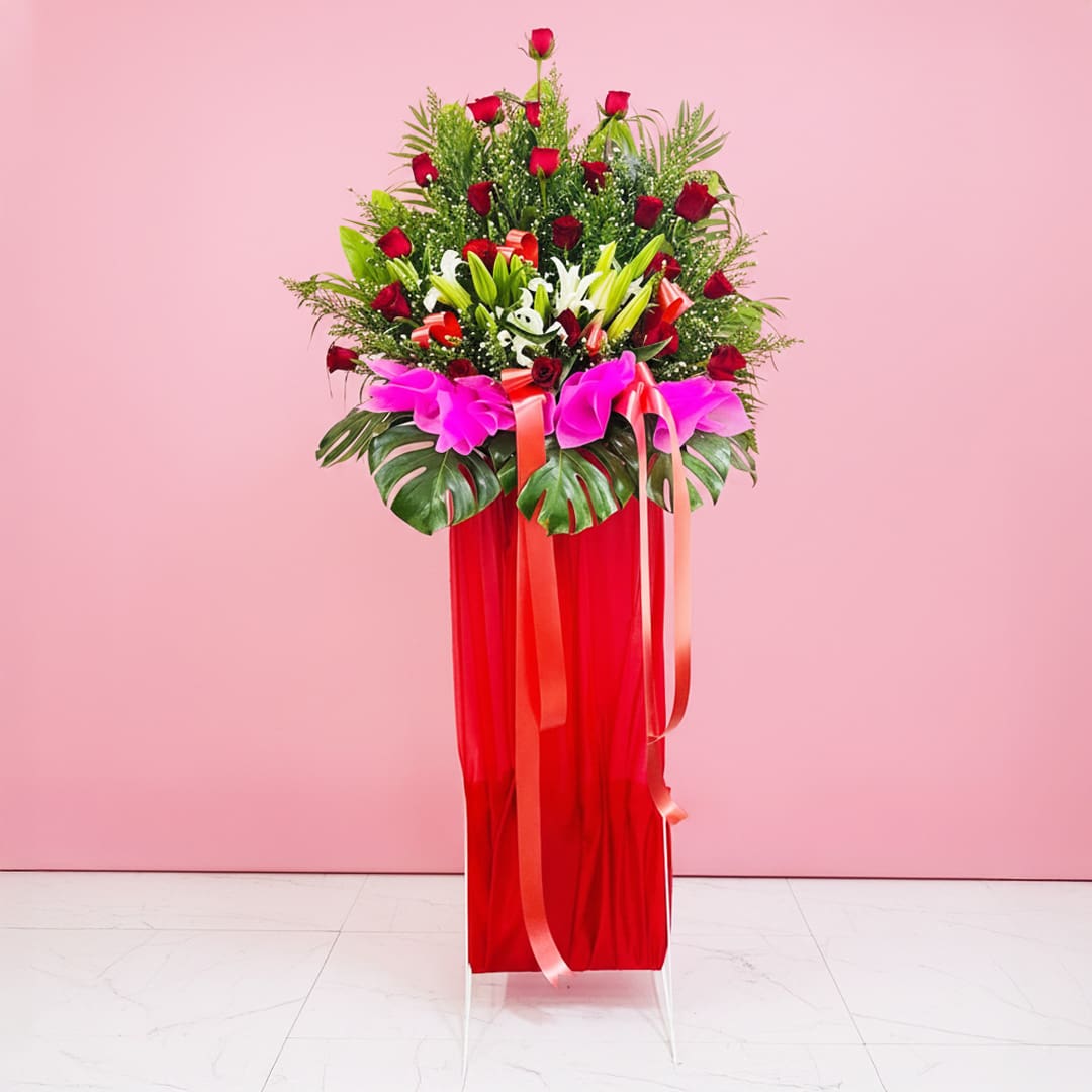 Flower Fusion Greeting Hub A red Grand Opening Stand filled with assorted flowers, including roses and lilies, adorned with a ribbon against a pink background.