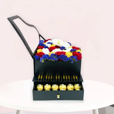 A black gift box surrounded by colorful flowers and an assortment of chocolates on a wooden table.