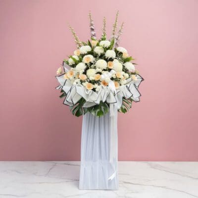 A tall condolence flower stand featuring white and cream blooms in a vase, set against a soft pink background.