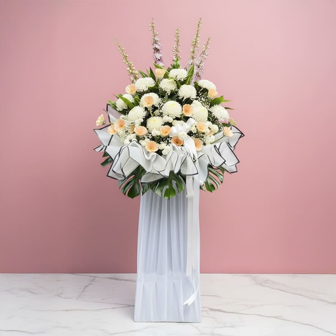 Heavenly Floral Whispers-01-240 A tall condolence flower stand featuring white and cream blooms in a vase, set against a soft pink background.