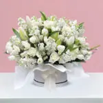 A round condolence box filled with white roses, tulips, lilies, and matthiola, elegantly arranged on a table.