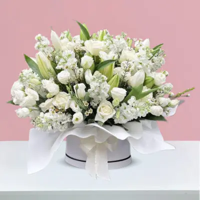 A round condolence box filled with white roses, tulips, lilies, and matthiola, elegantly arranged on a table.