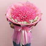 A woman stands holding a large bouquet of pink roses, showcasing their lush blooms and green foliage.