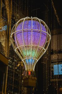 A large, illuminated hot air balloon sculpture hangs above a city street. The balloon is decorated with purple, blue, and yellow lights, creating a magical and festive atmosphere