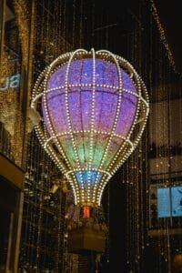 A large, illuminated hot air balloon sculpture hangs above a city street. The balloon is decorated with purple, blue, and yellow lights, creating a magical and festive atmosphere