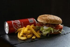 A delicious-looking fast food meal. The centerpiece is a sesame seed bun burger, overflowing with lettuce, tomato, and a crispy patty. Next to it, a pile of golden French fries and a can of Coca-Cola complete the picture.