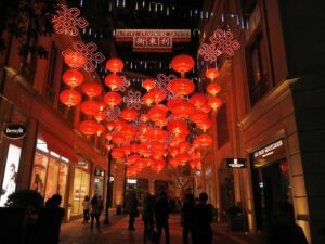 A bustling street scene in Hong Kong, likely during Chinese New Year. Colorful decorations adorn the storefronts, and people are walking along the sidewalks, some carrying shopping bags.