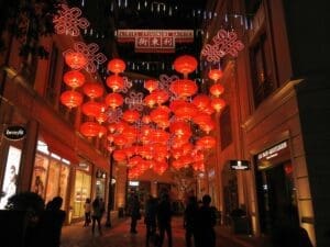 A bustling street scene in Hong Kong, likely during Chinese New Year. Colorful decorations adorn the storefronts, and people are walking along the sidewalks, some carrying shopping bags.