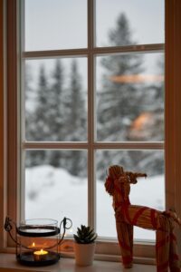 A cozy winter scene captured through a window. A straw goat decoration sits on a windowsill, with a lit candle and a small plant beside it. Outside, the window reveals a snowy landscape with tall trees in the distance.