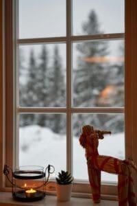 A cozy winter scene captured through a window. A straw goat decoration sits on a windowsill, with a lit candle and a small plant beside it. Outside, the window reveals a snowy landscape with tall trees in the distance.