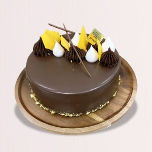 The image shows a round chocolate cake on a wooden plate. The cake is topped with chocolate shavings and white cream swirls. There are also some yellow decorations on the cake.