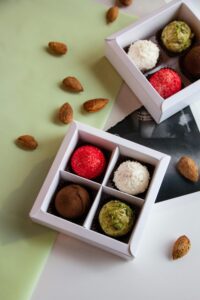 The image shows a variety of homemade truffles in white boxes. The truffles are in different colors, including red, white, brown, and green. There are also some almonds scattered around the boxes.