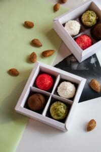 The image shows a variety of homemade truffles in white boxes. The truffles are in different colors, including red, white, brown, and green. There are also some almonds scattered around the boxes.