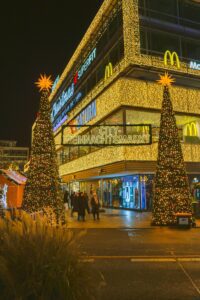 Two large, illuminated Christmas trees stand in front of a building adorned with golden lights and the words "Christmas" and "Weihnachtsmarkt" (Christmas Market) displayed prominently.