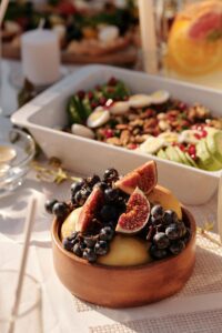 he image shows a bowl of fresh fruit on a table. The bowl contains slices of figs and grapes. In the background, there is a salad in a white dish.