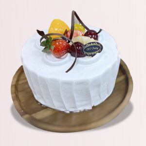 The image shows a round cake with white frosting and a variety of colorful fruits on top. The cake is on a wooden plate.