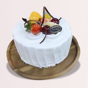 The image shows a round cake with white frosting and a variety of colorful fruits on top. The cake is on a wooden plate.