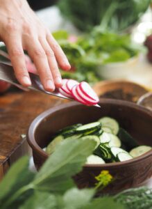 A close-up shot of a hand slicing a radish on a cutting board. The freshly sliced radish rounds are falling onto a bowl filled with sliced cucumbers