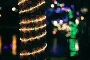 A tree trunk wrapped with colorful LED lights. The lights create a warm and festive glow against the dark background. Other lights can be seen in the background, creating a bokeh effect. The image evokes a sense of holiday cheer and celebration.